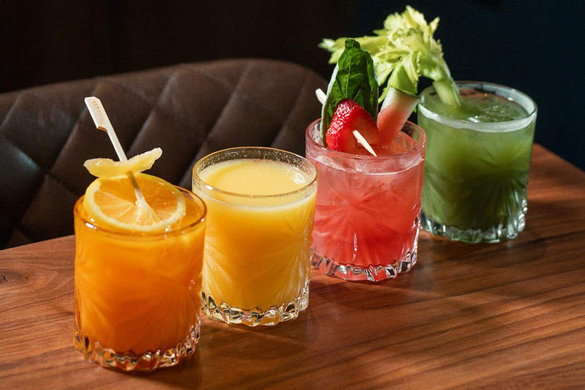 Four tumbler glasses filled with juices of various colors, orange, yellow, red and green, with accompanying garnishes, all sitting on a wooden tabletop with a brown leather cross stitched chair in the background