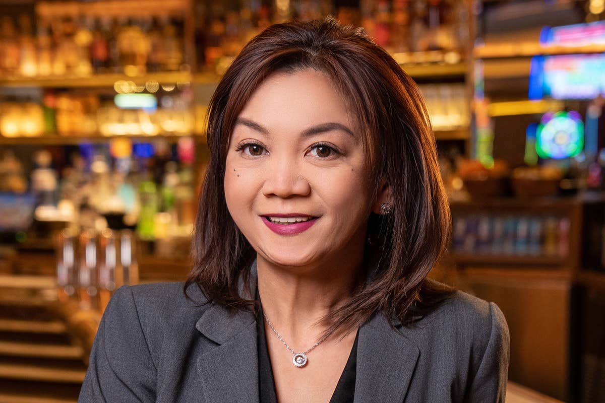 A woman with shoulder-length hair and a gray blazer smiles in front of a bar background.