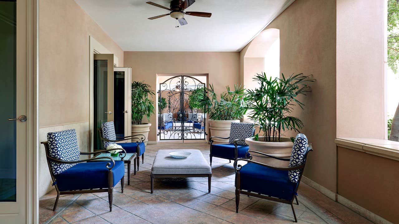 Outdoor patio with blue cushioned chairs, a central ottoman, potted plants, ceiling fan, and iron gate in the background.