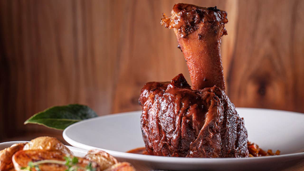 A plated braised lamb shank served with roasted vegetables, garnished with a bay leaf on a wooden background.