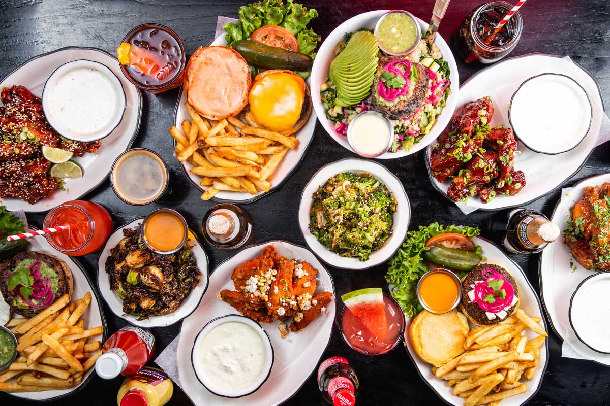 An overhead view of various dishes including burgers, fries, wings, salads, and drinks on a dark tabletop.