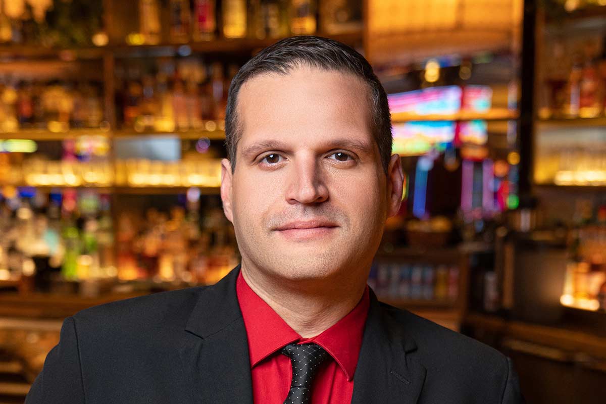 A man in a black suit and red shirt stands in front of a well-lit bar with bottles and glasses in the background.