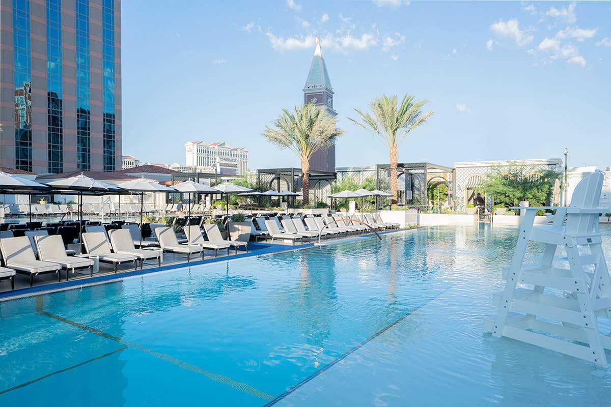 Rooftop pool area with lounge chairs, umbrellas, palm trees, and a distant clock tower under a clear blue sky.