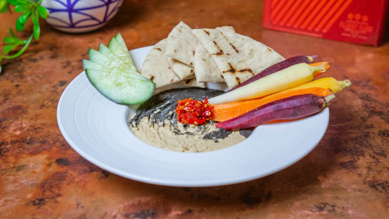 A white plate with hummus, sliced cucumber, pita bread, colorful carrots, and a red pepper garnish.