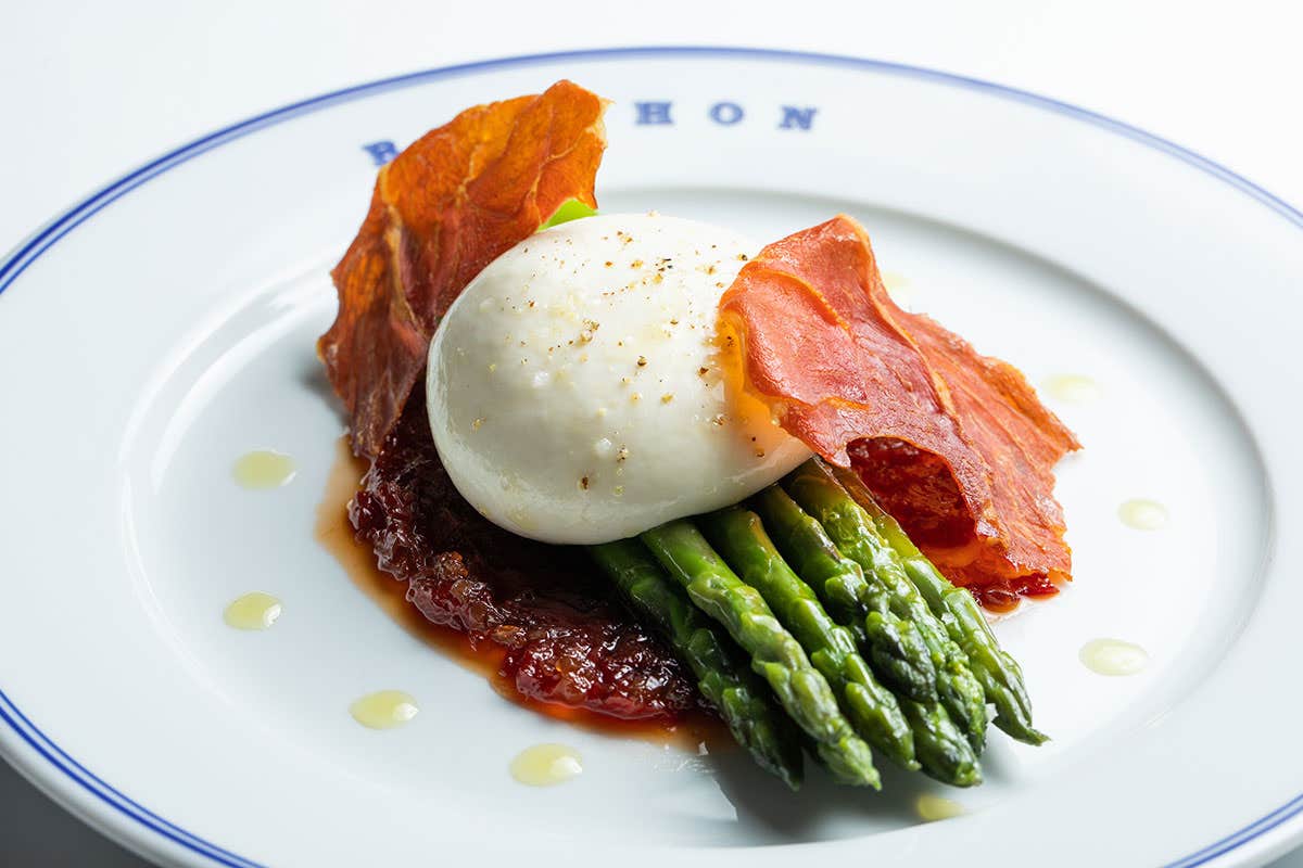 A gourmet plate featuring burrata cheese, crispy prosciutto, asparagus, and sauce on a white plate garnished with oil drops.