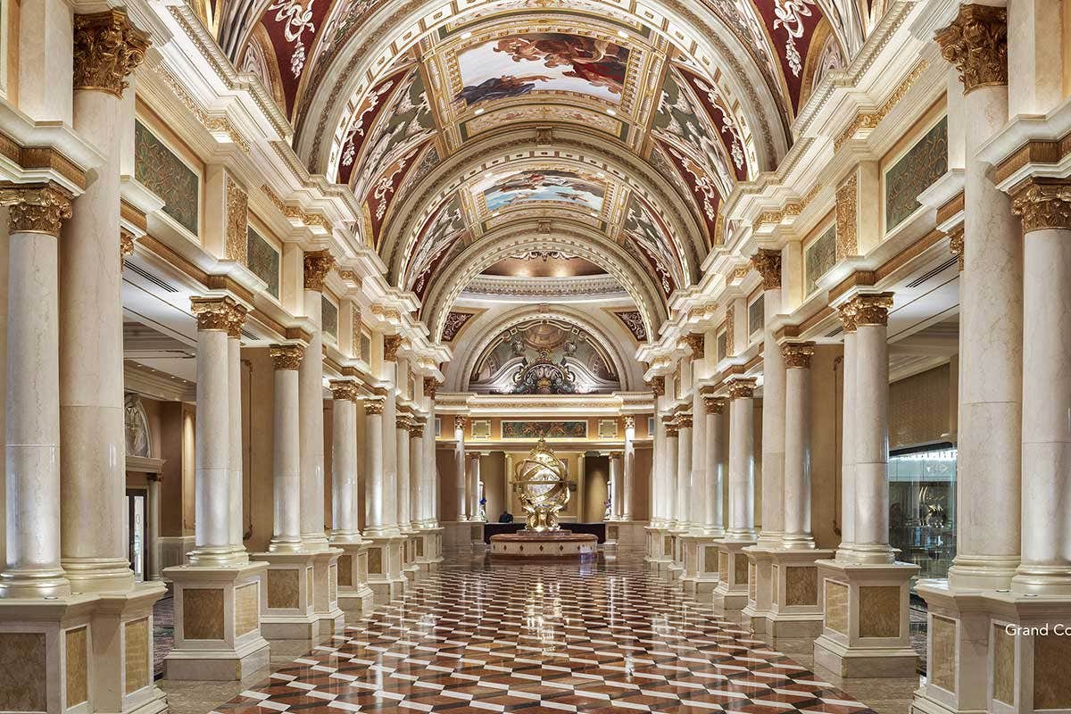 Opulent corridor with vaulted ceilings, ornate columns, frescoes, and checkered floor tiles leading to a large decorative globe.