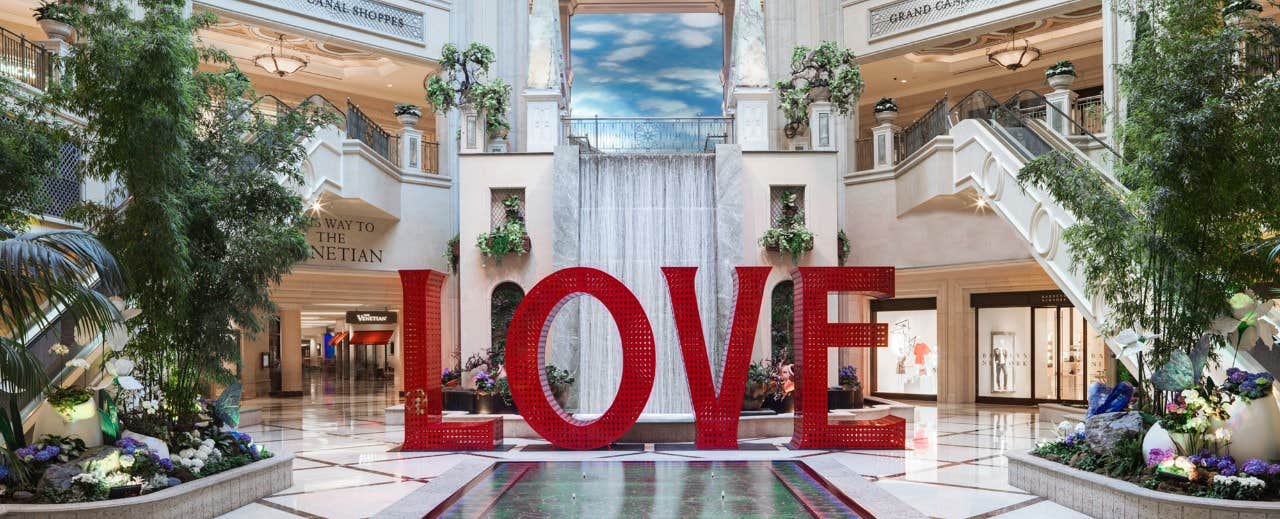 Large red "LOVE" sculpture in front of a indoor waterfall at a grand shopping mall entrance with elegant decor and greenery.