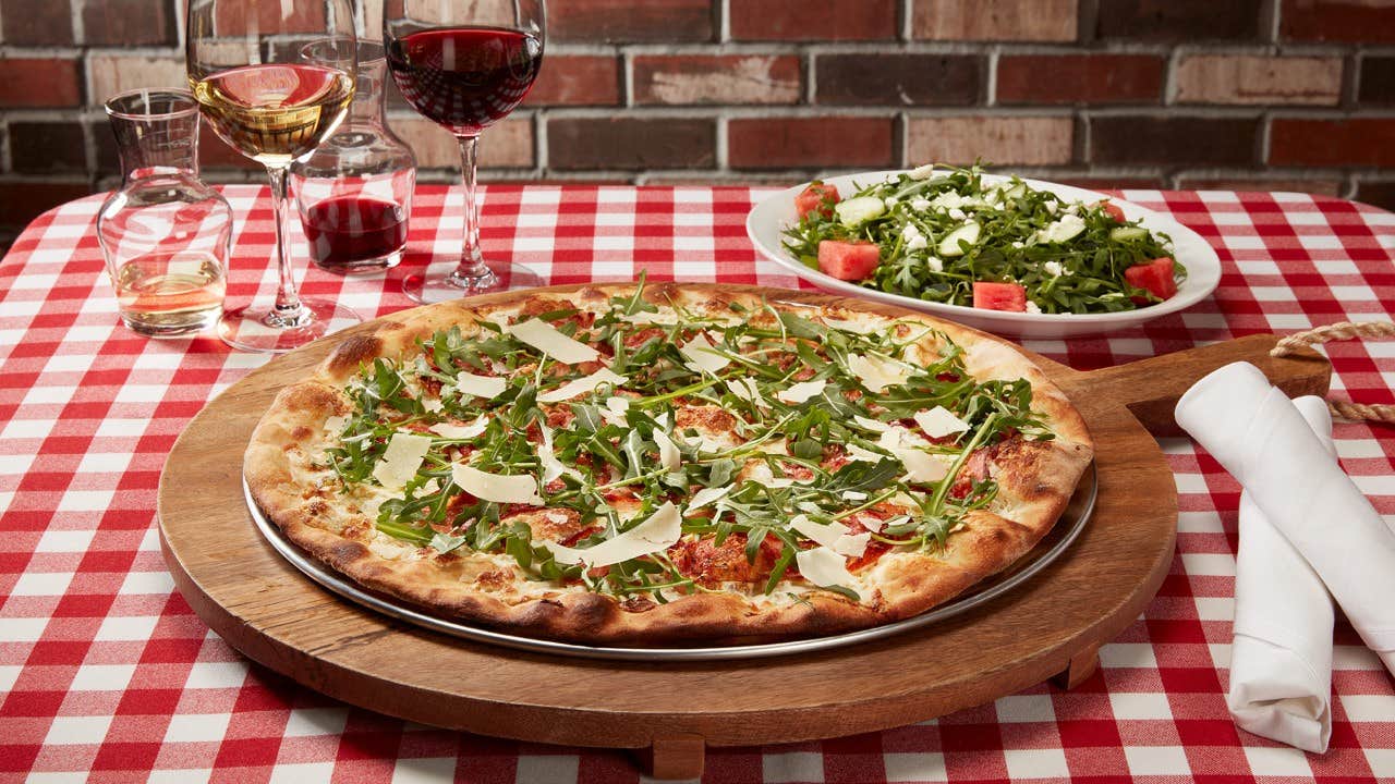 A pizza topped with arugula and cheese on a wooden board, with wine glasses and a salad on a red-checkered tablecloth.