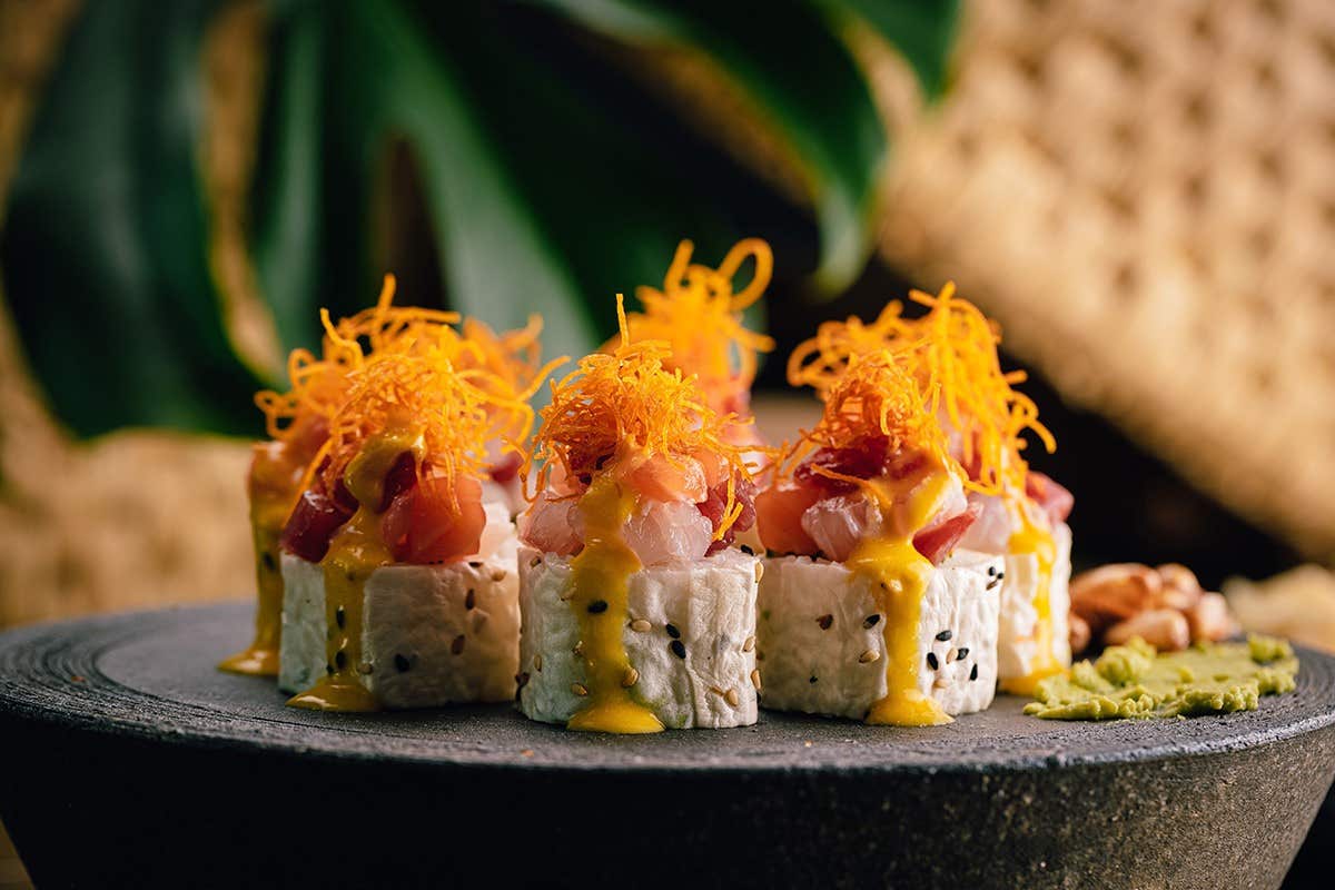 Close-up of gourmet sushi rolls topped with shredded orange garnish and yellow sauce, served on a dark stone plate.