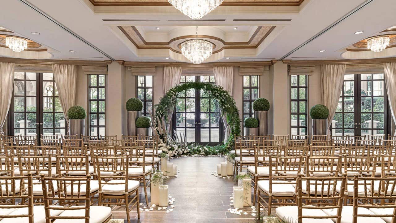 Elegant wedding venue with chandeliers, greenery, white flowers, and rows of chairs facing a floral arch in front of windows.