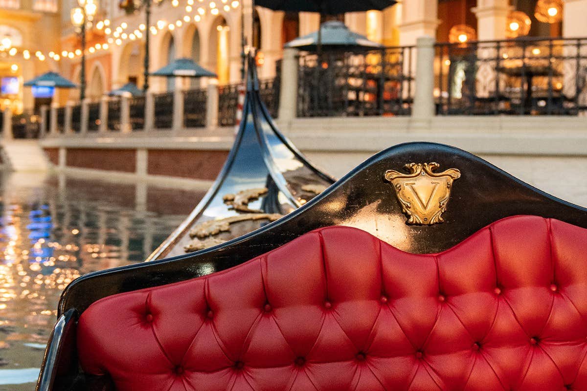 Red tufted seat in a gondola with a "V" emblem, floating in a canal with ornate buildings and string lights in the background.