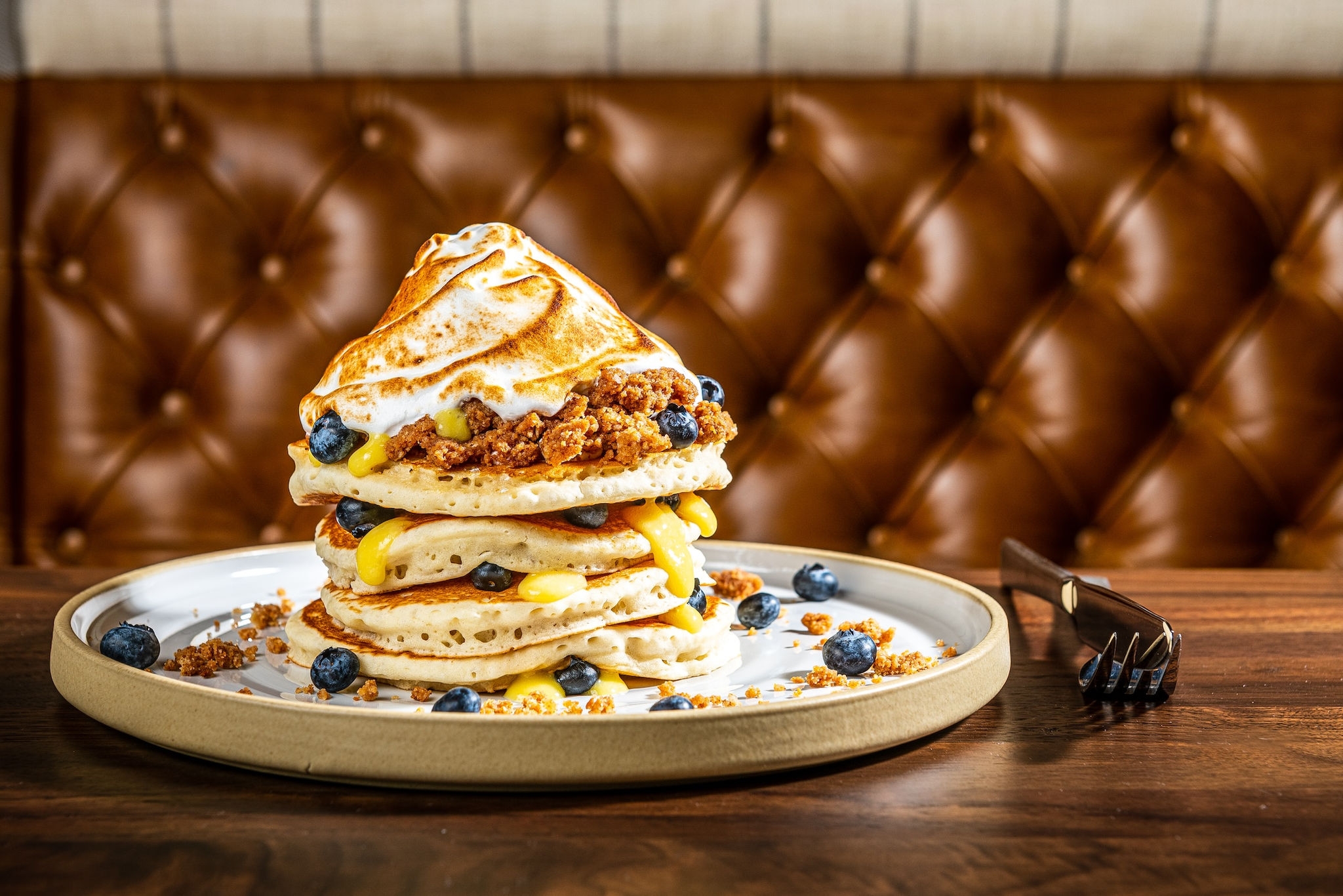 A tall stack of pancakes with crumbled toppings, blueberries, and whipped cream on a plate, set against a tufted leather booth.