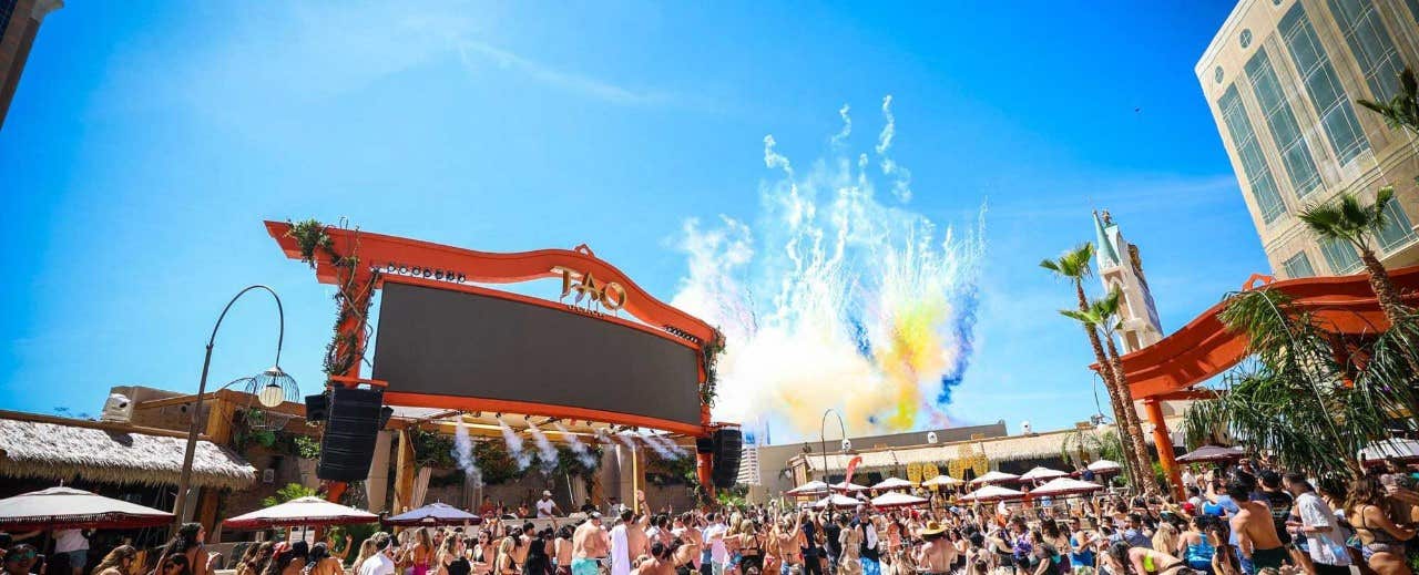 Outdoor pool party with a large screen, DJ stage, crowds of people, and colorful smoke effects under a bright blue sky.