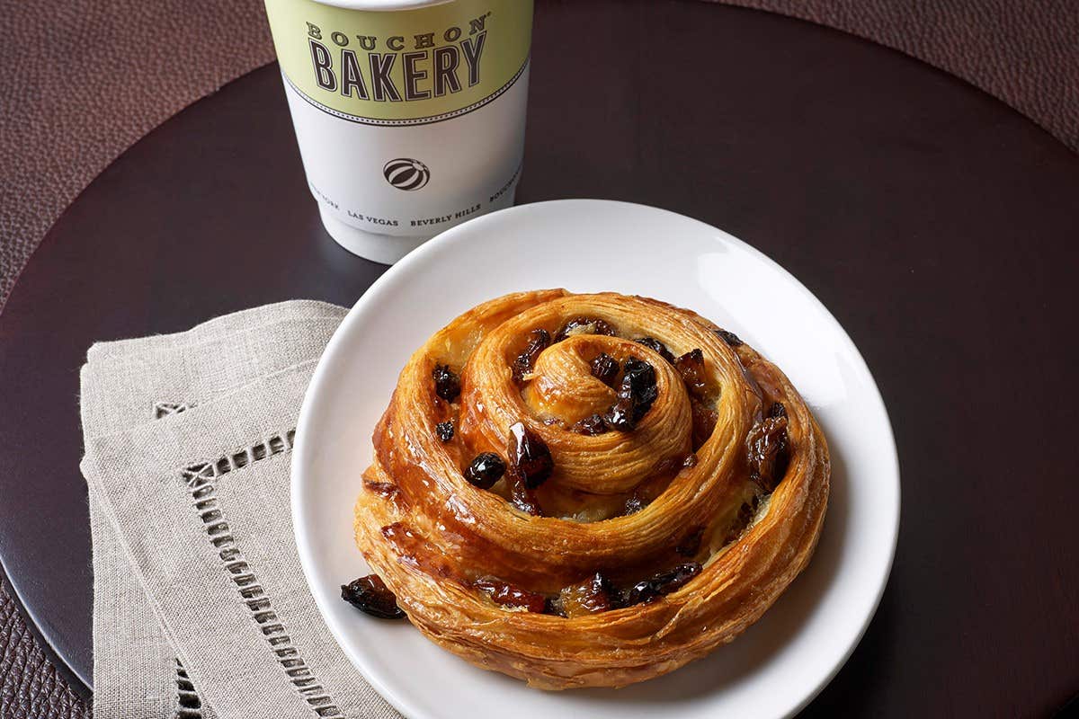 Pastry swirl with raisins on a white plate next to a Bouchon Bakery coffee cup and a beige napkin.