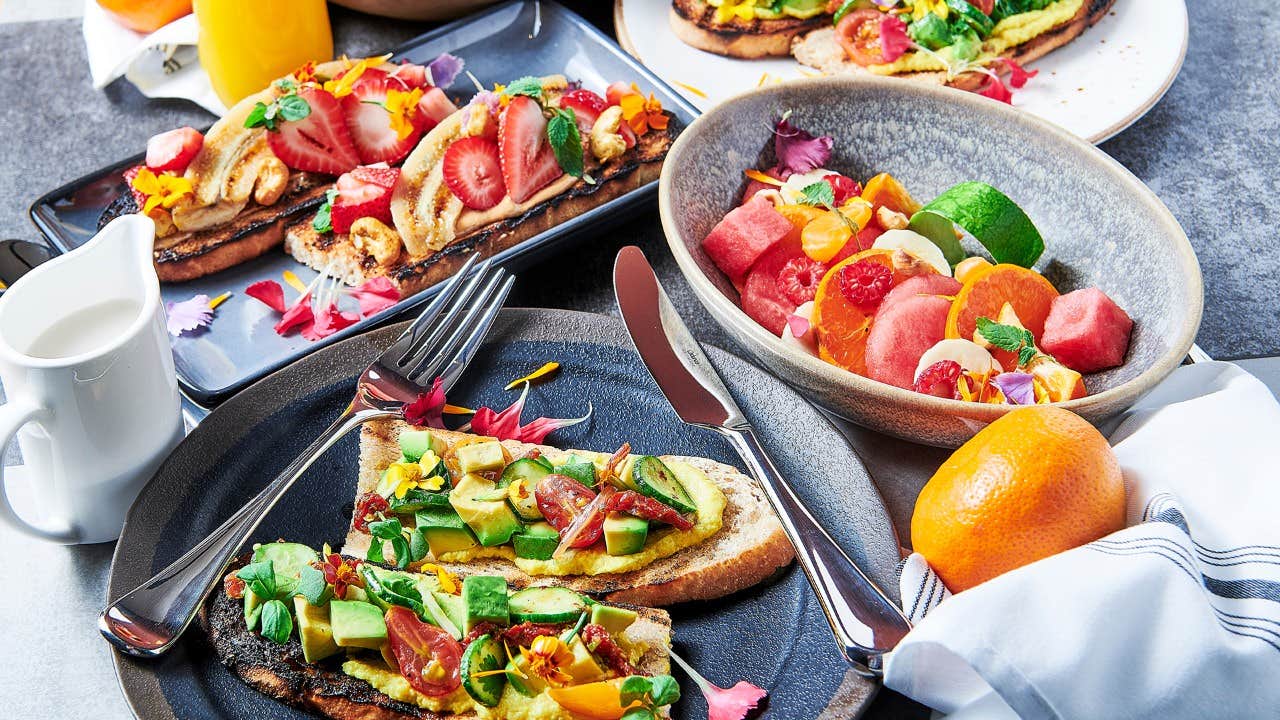 A vibrant brunch spread featuring avocado toasts, fruit salad, an orange, and a small pitcher of milk.