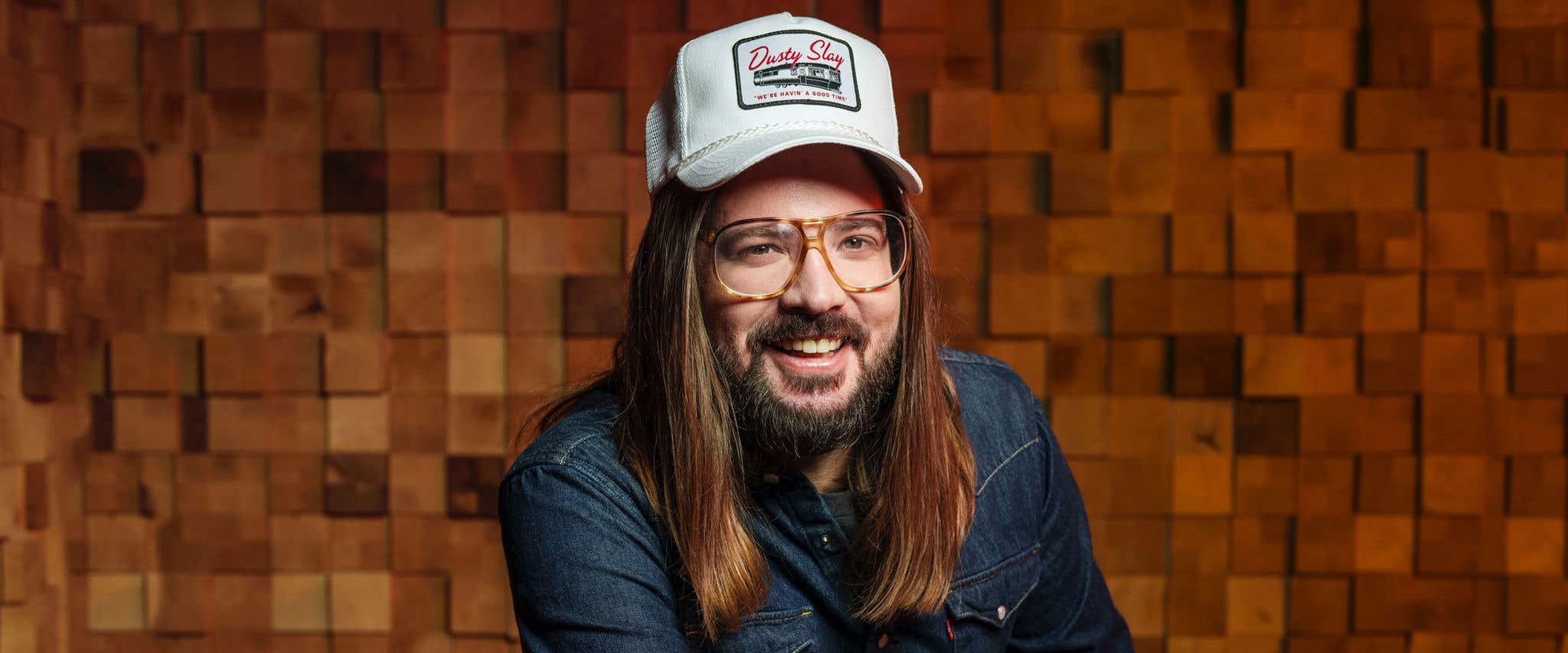 Dusty Slay with long hair and glasses, wearing a denim shirt and a white cap, smiles in front of a wooden textured background.