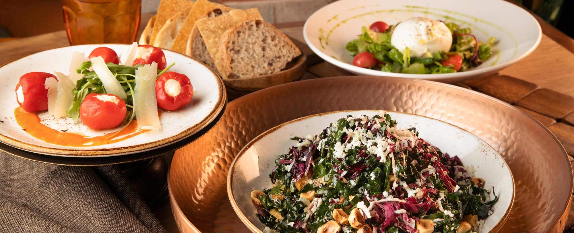 Three plates of food on a table: a salad with greens and nuts, stuffed tomatoes with greens, and a dish with bread slices.