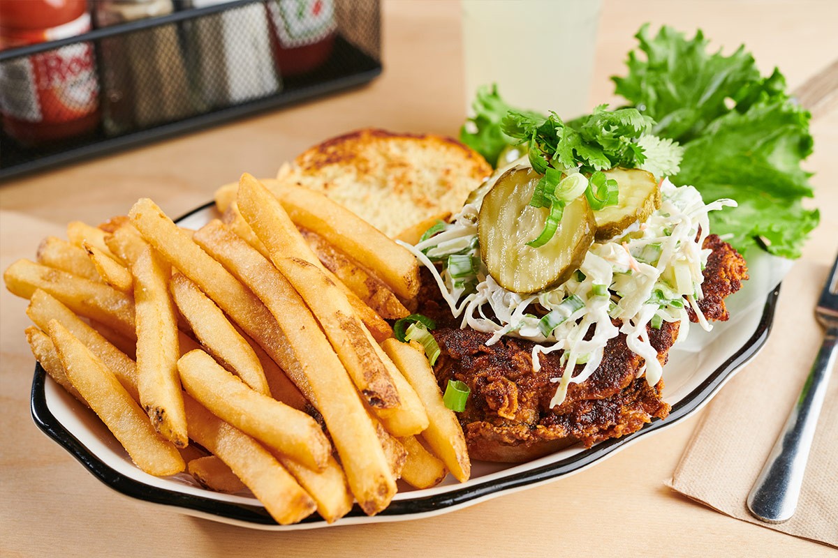 Plate of crispy French fries and a chicken sandwich topped with coleslaw, pickles, lettuce, and herbs on a table.