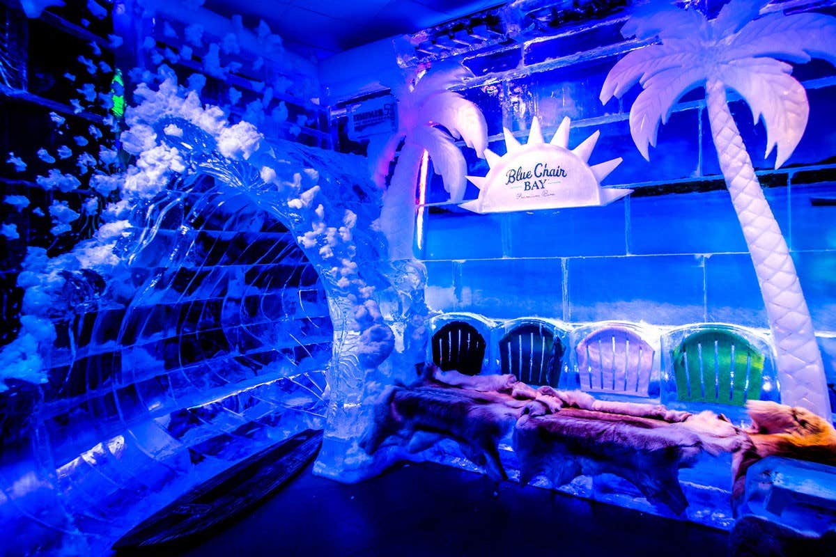 A vibrant ice bar with ice sculptures of palm trees, a wave tunnel, and seating with fur throws under purple lighting.