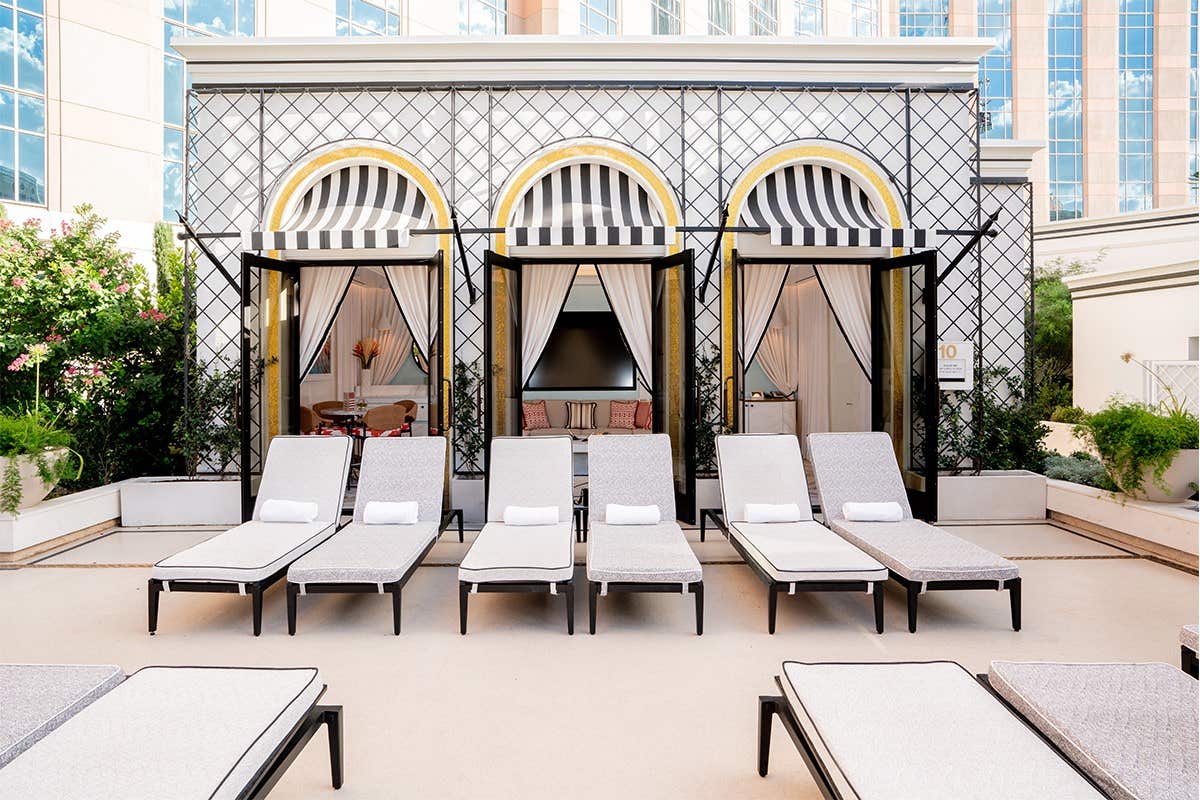 Poolside cabanas with black-and-white striped awnings and lounge chairs in front, with a building in the background.