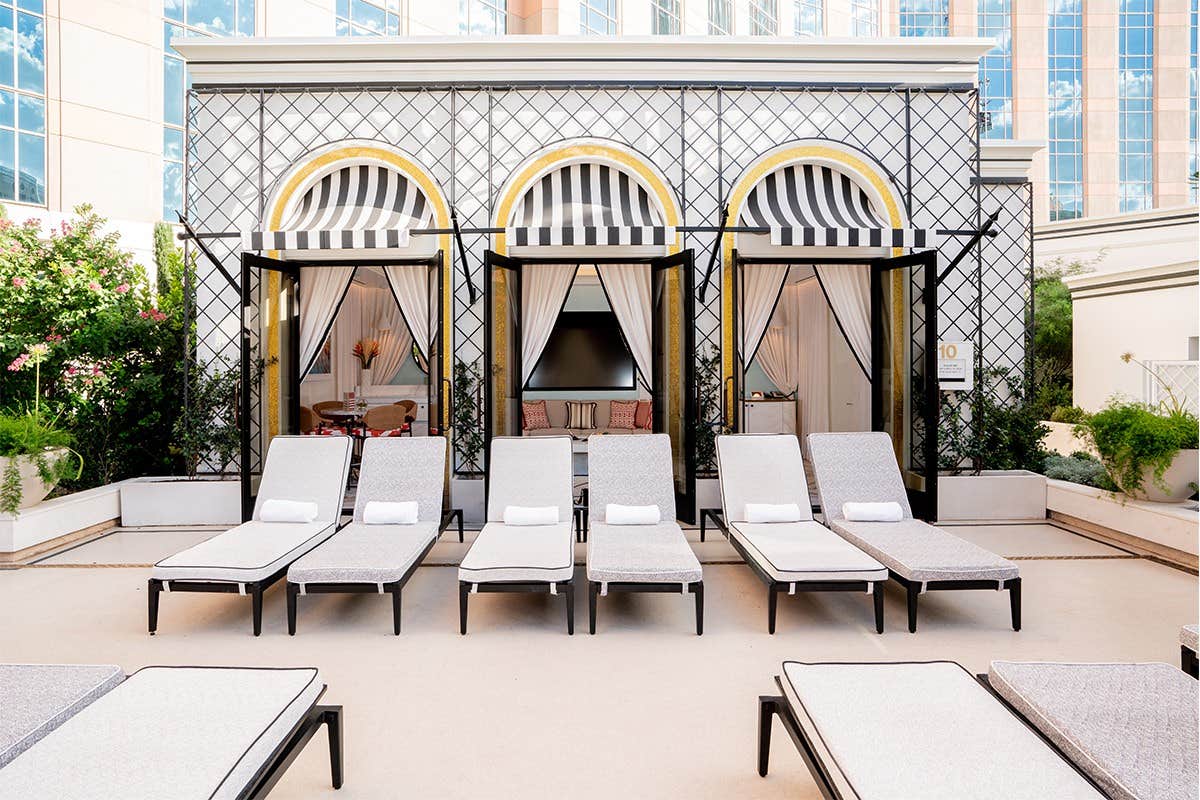 Poolside cabanas with black-and-white striped awnings and lounge chairs in front, with a building in the background.