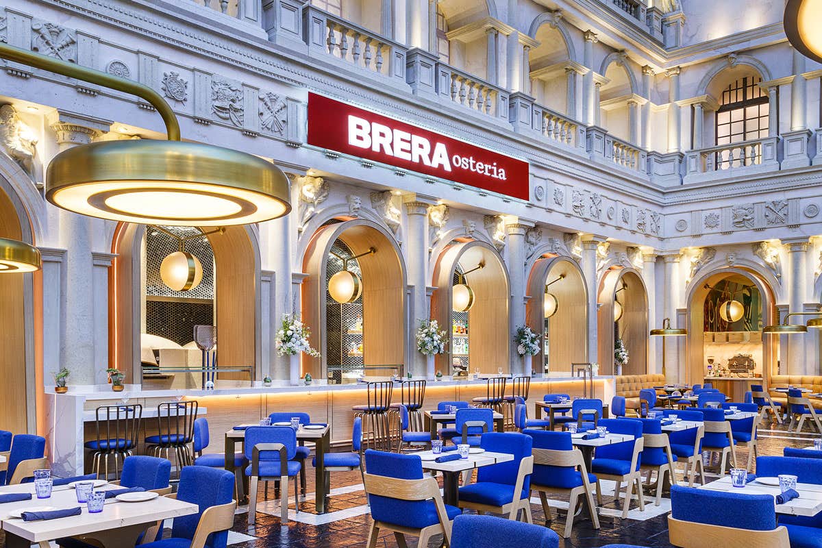 Modern restaurant with blue and white seating, large gold hanging lights, and an illuminated "BRERA osteria" sign inside.