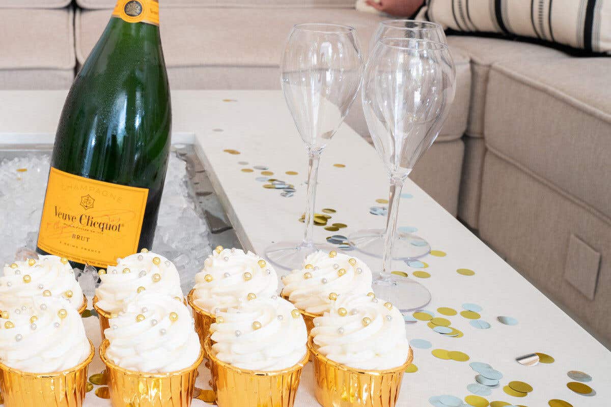 A bottle of champagne surrounded by six cupcakes in gold wrappers and two empty champagne flutes on a confetti-covered table.