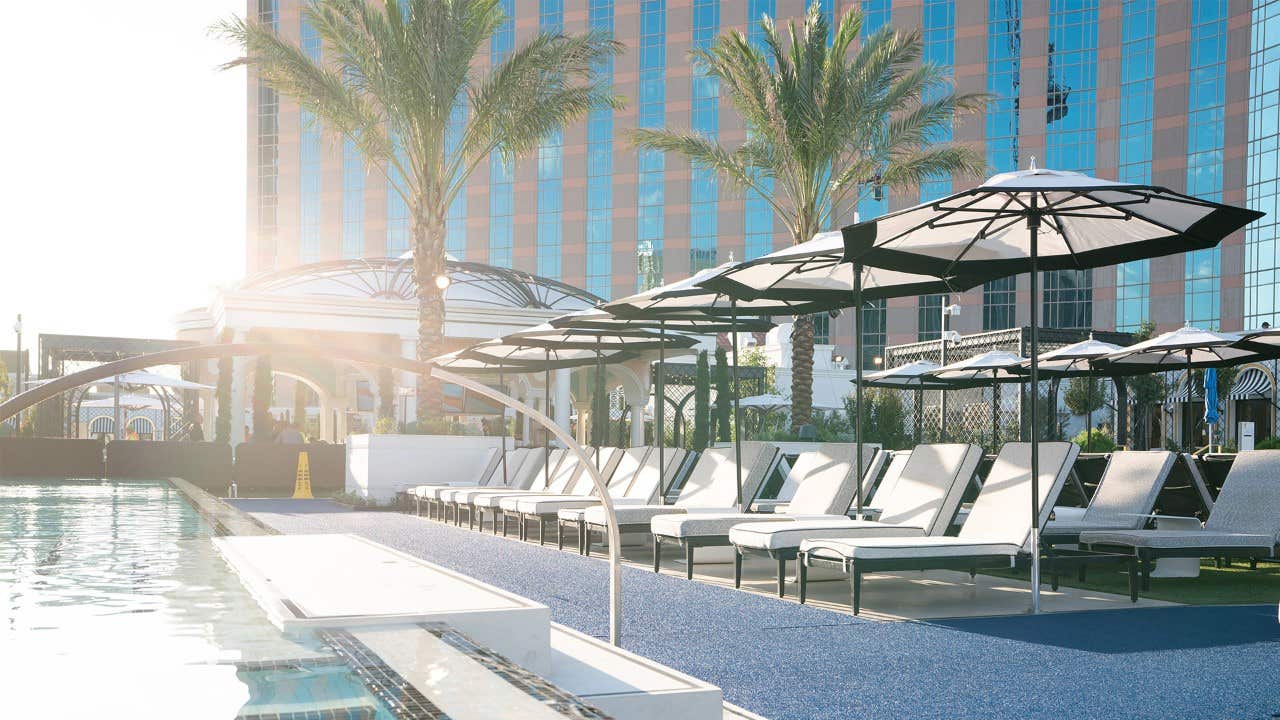 A serene poolside area with sun loungers, umbrellas, palm trees, and a modern building in the background at sunset.