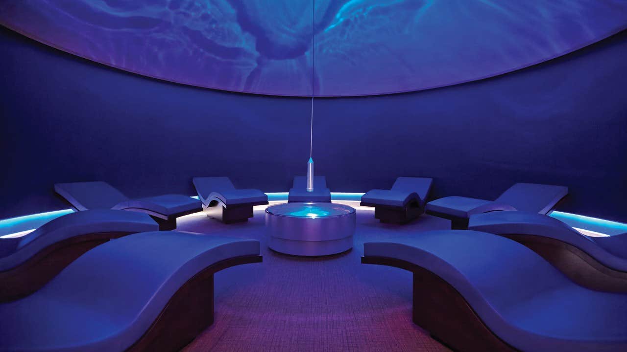 A dimly lit relaxation room with blue ambiance, featuring curved lounge chairs around a central water fixture.