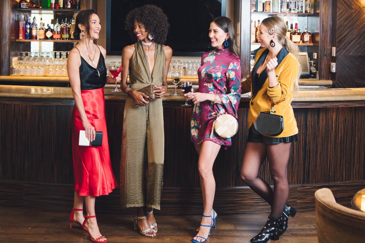 Four women dressed fashionably stand conversing in a stylish bar setting, smiling and holding drinks.