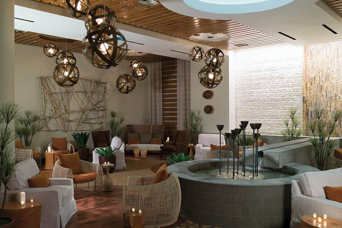 A modern lounge with wicker and upholstered seating, decorative lighting, plants, and a central fountain feature.