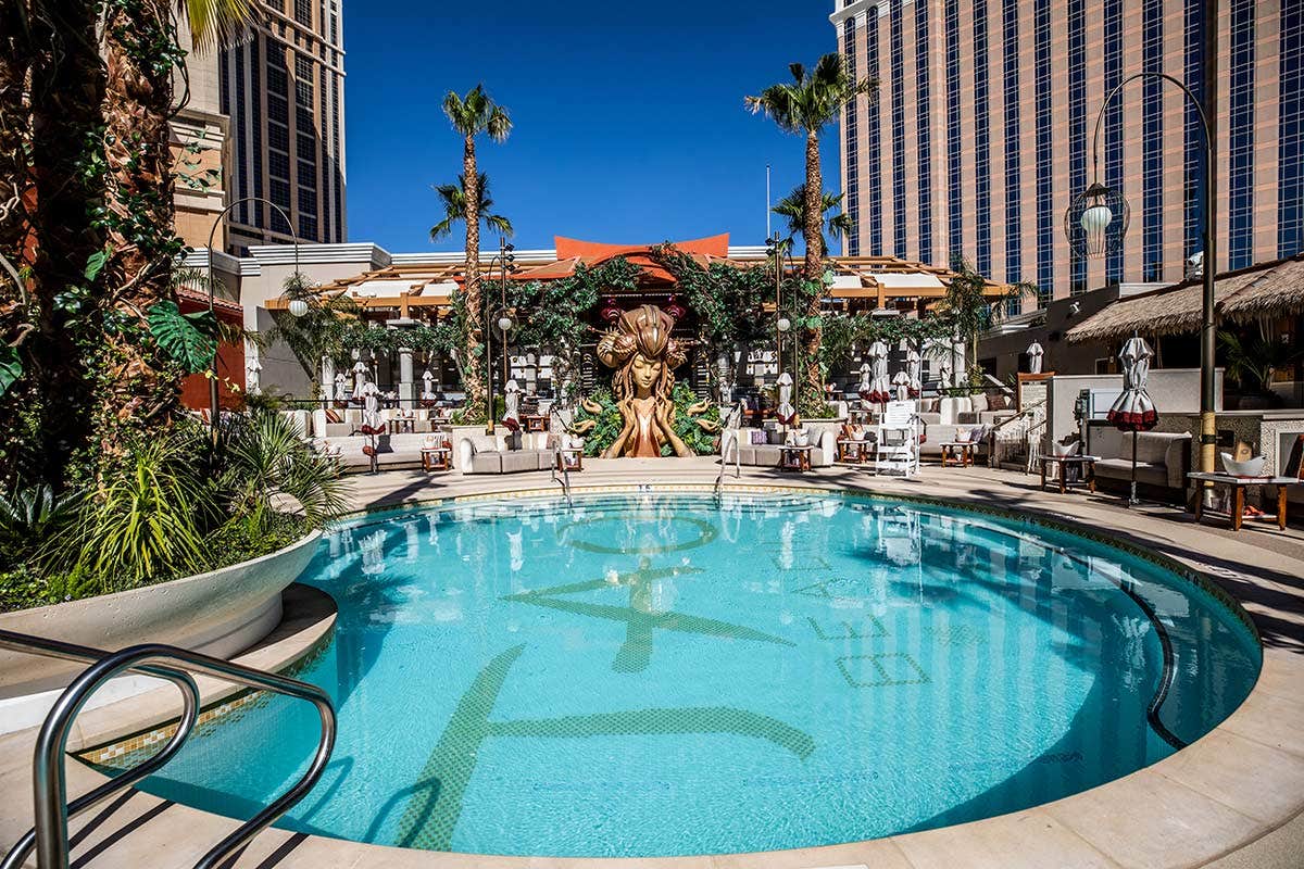 Outdoor resort pool surrounded by palm trees, lounge chairs, and cabanas, with buildings and blue sky in the background.