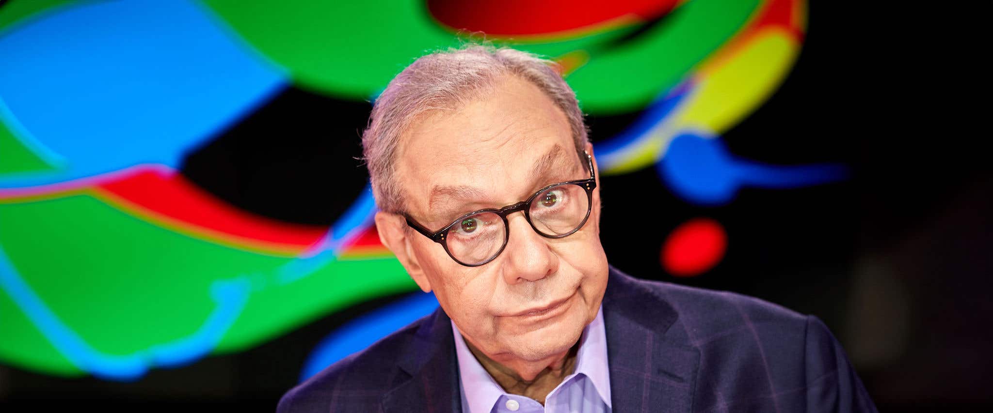 Lewis Black in a blue suit jacket in front of a colorful background