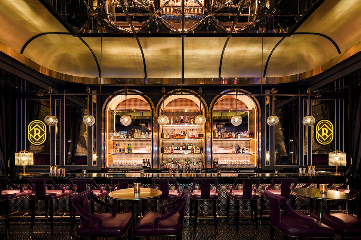 Elegant bar interior with arched mirrors, gold accents, lit shelves of bottles, purple seating, and ambient lighting.