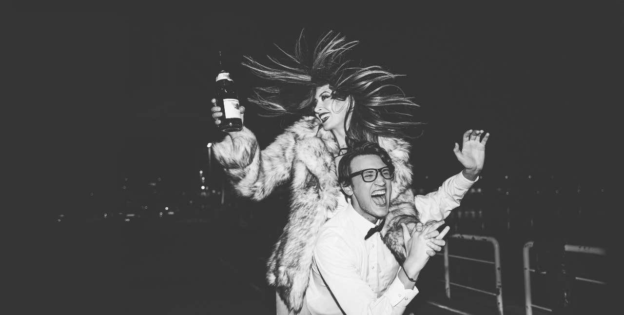 A woman holding a bottle and a man in glasses laugh and celebrate at night in black and white.