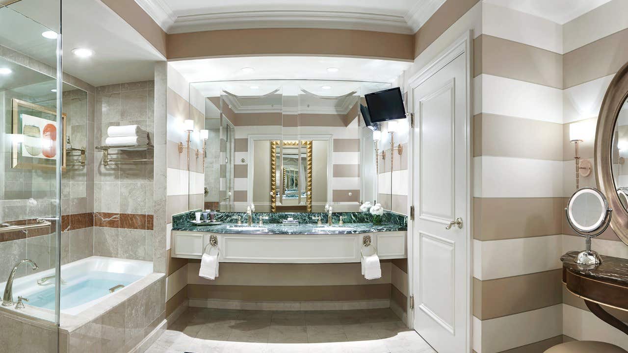 Modern bathroom with striped walls, double sink vanity, bathtub, and mounted TV above a door. Large mirror and lights.