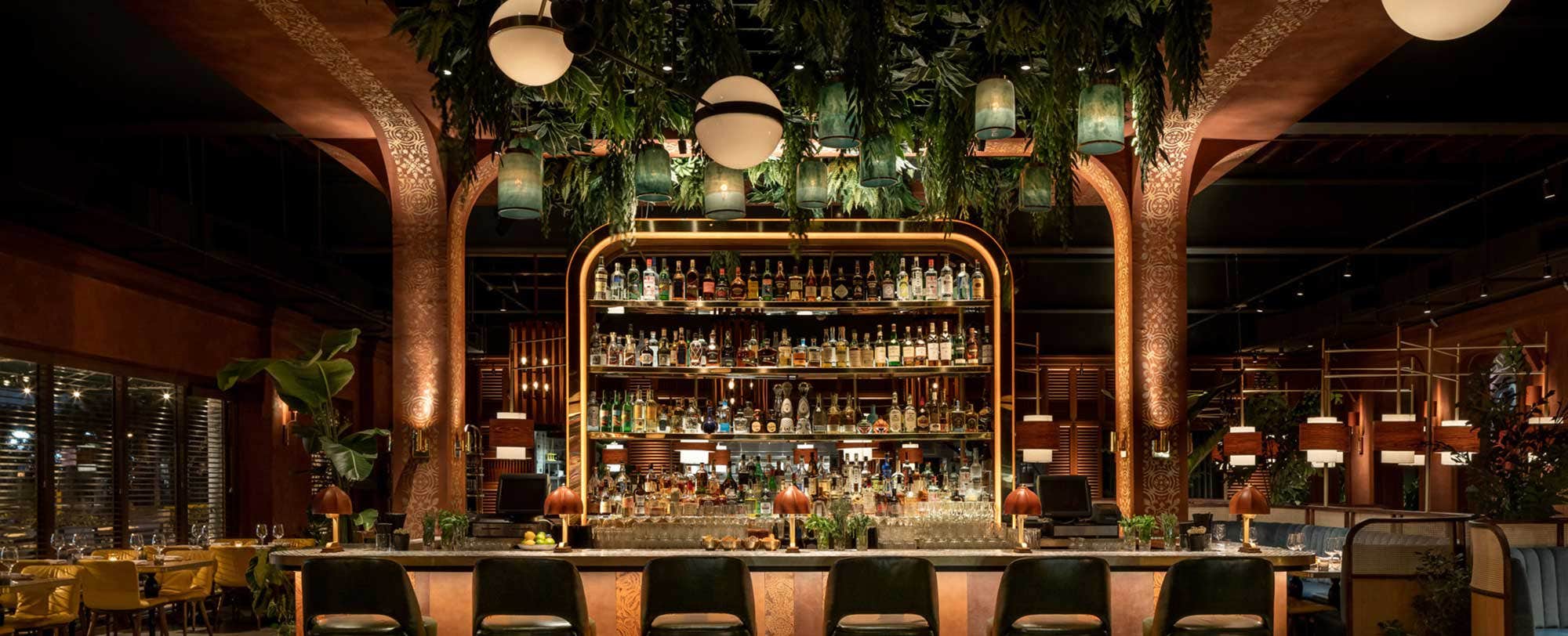 A luxurious bar with green chairs, hanging plants, and an array of bottles on backlit shelves in an elegant, dimly lit setting.