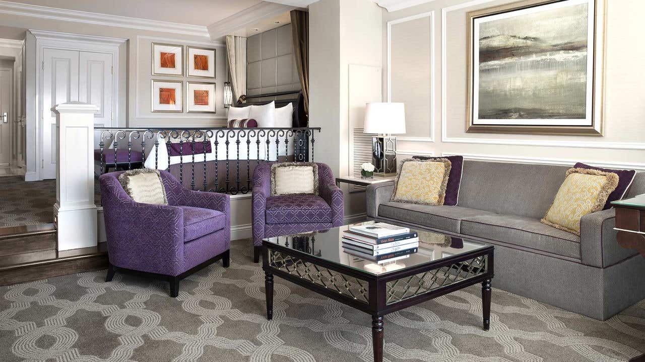 A stylish living area with purple chairs, a gray sofa, a glass coffee table, and a bed in a partially separated space.