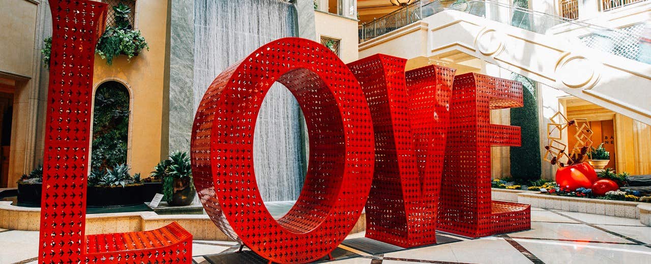 Large red "LOVE" sculpture indoors near a fountain and greenery, with stairs and decorative features in the background.