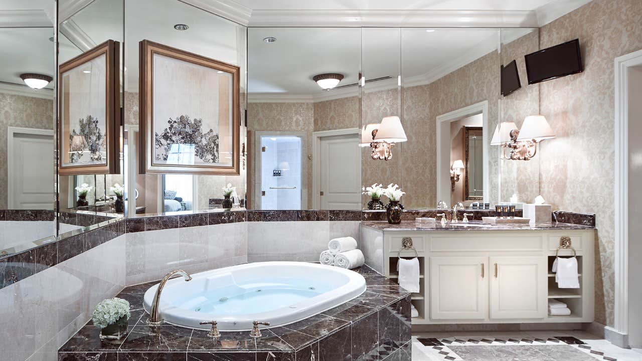 Elegant bathroom with a jacuzzi tub, vanity, and mirrors. Beige wallpaper, dark countertops, and stylish lighting fixtures.