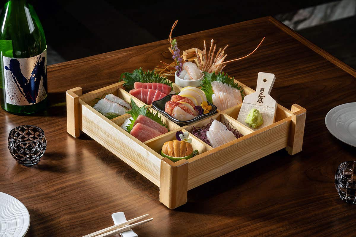 A wooden tray with assorted sashimi and sushi pieces, a bottle of sake, and two glasses on a wooden table.