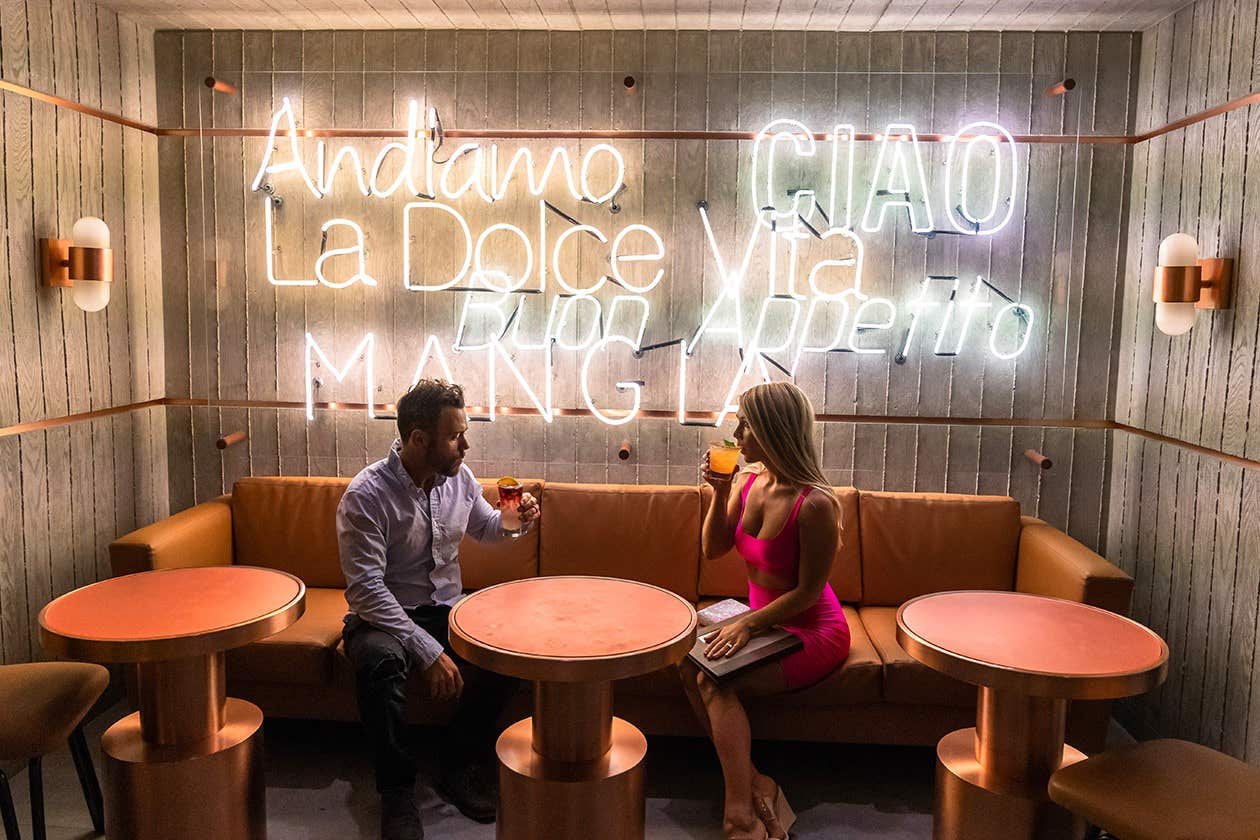 A man and woman drinking cocktails in a cozy lounge with neon signs and a brown leather sofa.