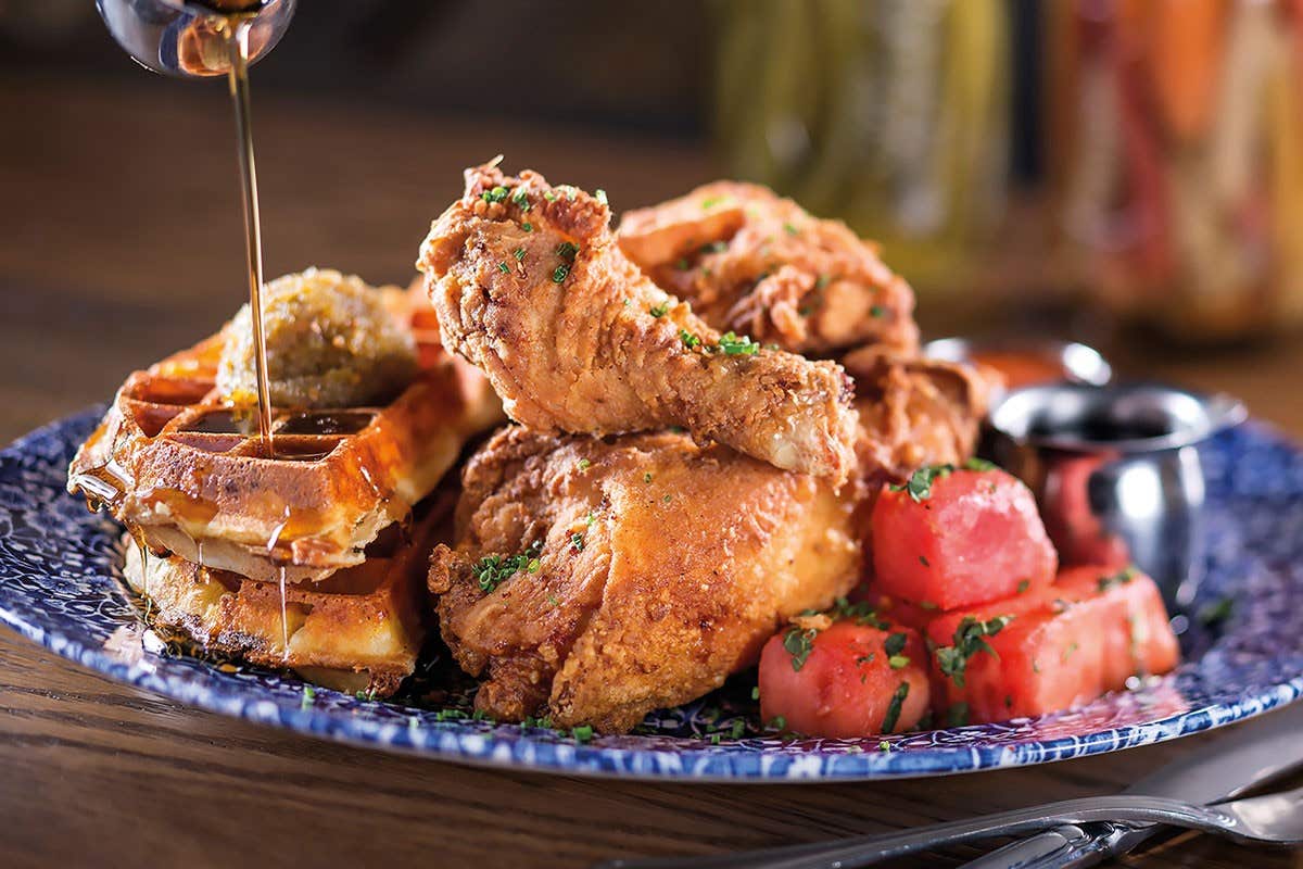 A plate of fried chicken with waffles, syrup being poured, served with watermelon chunks and syrup cups.