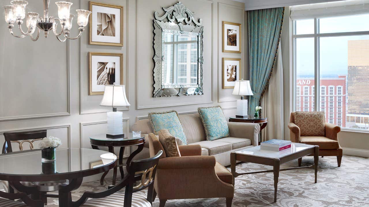 Elegant hotel suite with sofa, armchairs, coffee table, mirror, wall art, chandelier, dining table, and city view window.