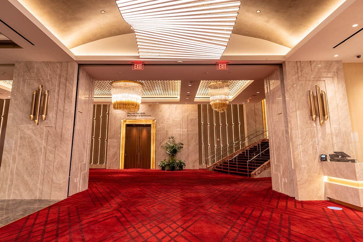 Hotel lobby with marble walls, red carpet, elegant chandeliers, and a staircase; mirrored ceiling lights.