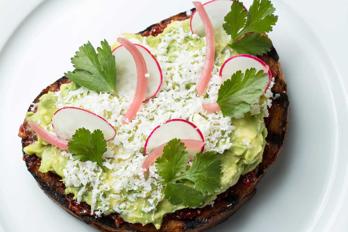 Toast topped with mashed avocado, radish slices, grated cheese, and cilantro leaves on a white plate.