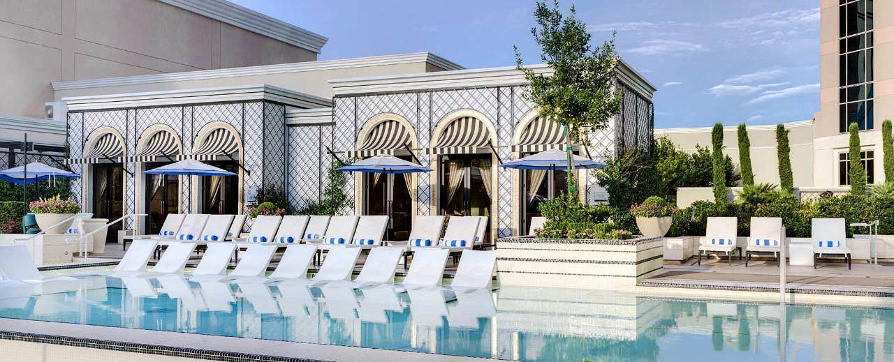 A luxurious pool area with white lounge chairs, blue umbrellas, and elegant cabanas in the background.