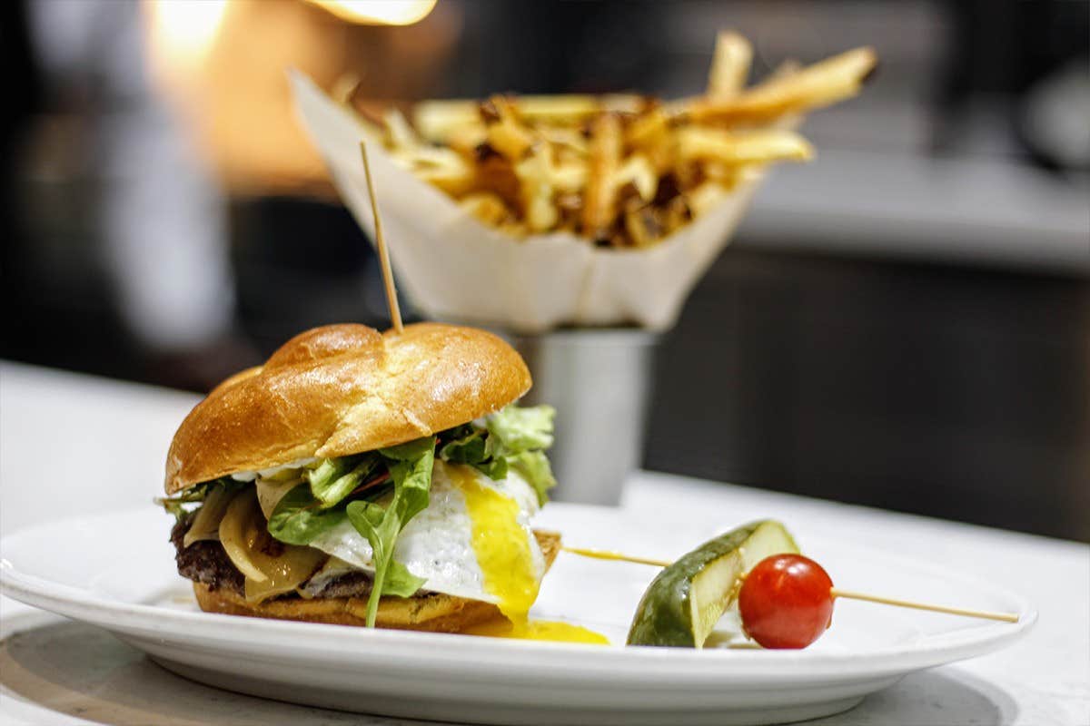 A burger with lettuce, egg, and cheese on a white plate served with fries, a pickle, and a cherry tomato.